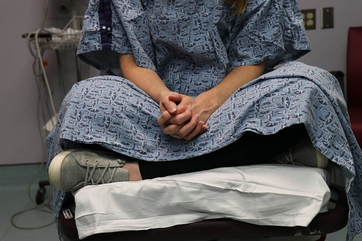 The lower half of an unidentifiable patient is shown wearing a hospital gown and sitting on a table in a hospital room waiting to be examined by a sexual assault nurse examiner (sane).jpg