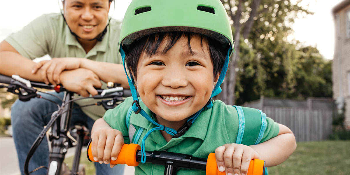 A young boy wearing a helmet poses smiling at the camera while on his bike.  His father is doing the same in the background.
