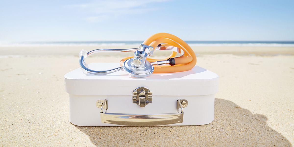 Stethoscope and suitcase on beach by the sea, symbol for travel pharmacy/ first aid kit