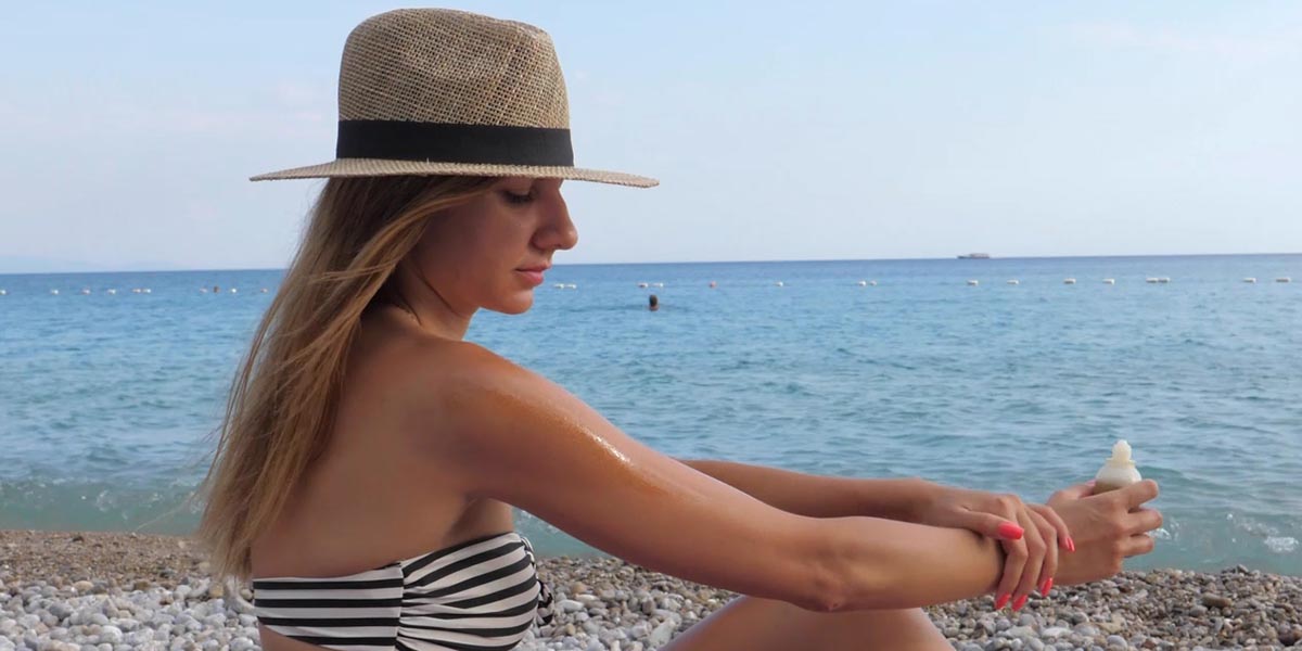 Woman wearing a hat and applying sunscreen, while sitting on a beach