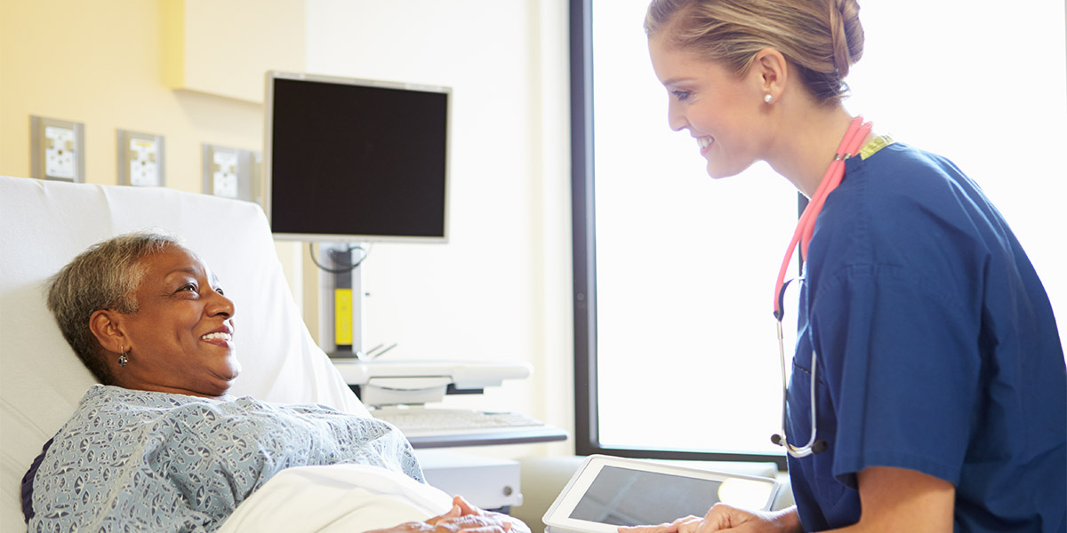 Nurse With Digital Tablet Talks To Woman In Hospital Bed