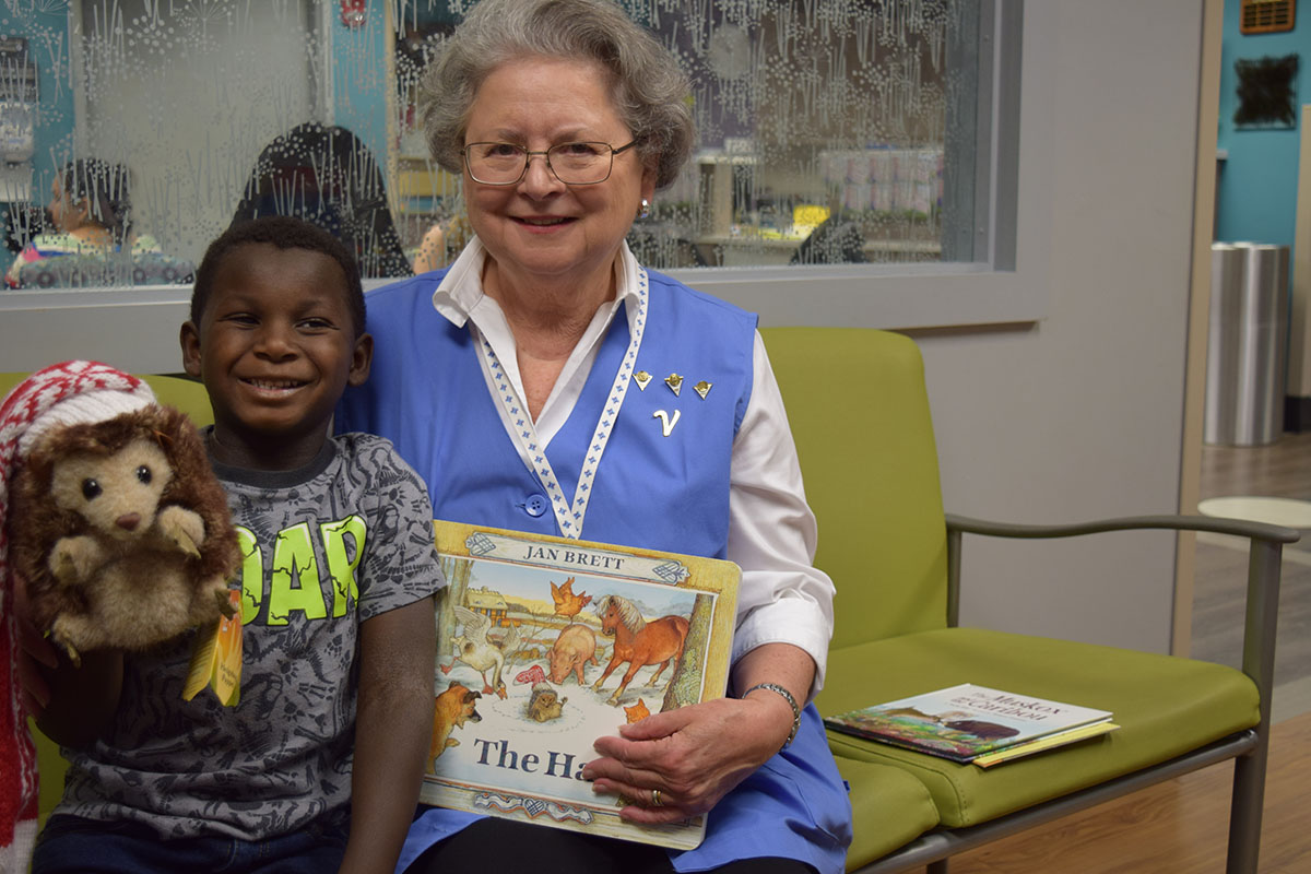 A mature volunteer holds a book posing with a young boy she reads to while he waits for his pediatric rehab program appointment smiling and holding a teddy bear beside her.jpg