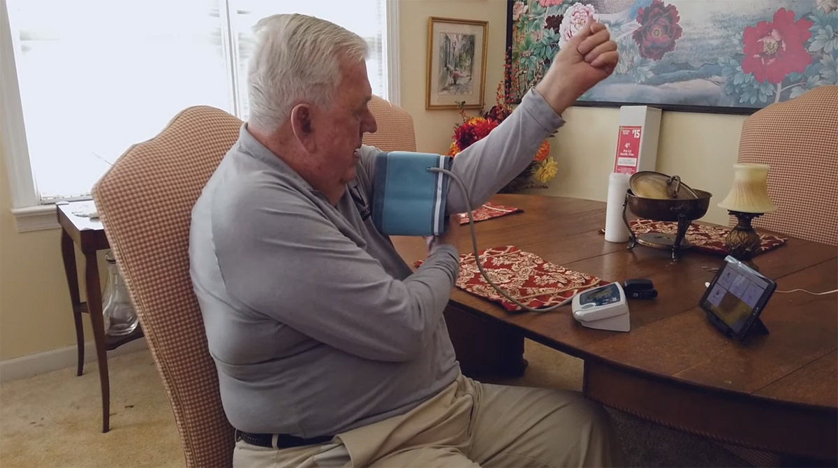 An elderly man sits at a dining room table with a blood pressure cuff to track his own health